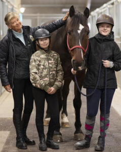 Äldre kvinna och två barn står i stallgång med brun häst. Smedstad Ridsportcenter